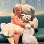 Child in fairy costume cuddling giant gray teddy bear on couch