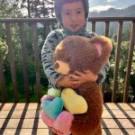 Boy in bear hat holding brown teddy bear with colorful hearts outdoors
